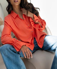 Coral LA TROYES blouse by PLEIN PUBLIQUE, shown worn with one hand on the collar and the other resting on her leg.