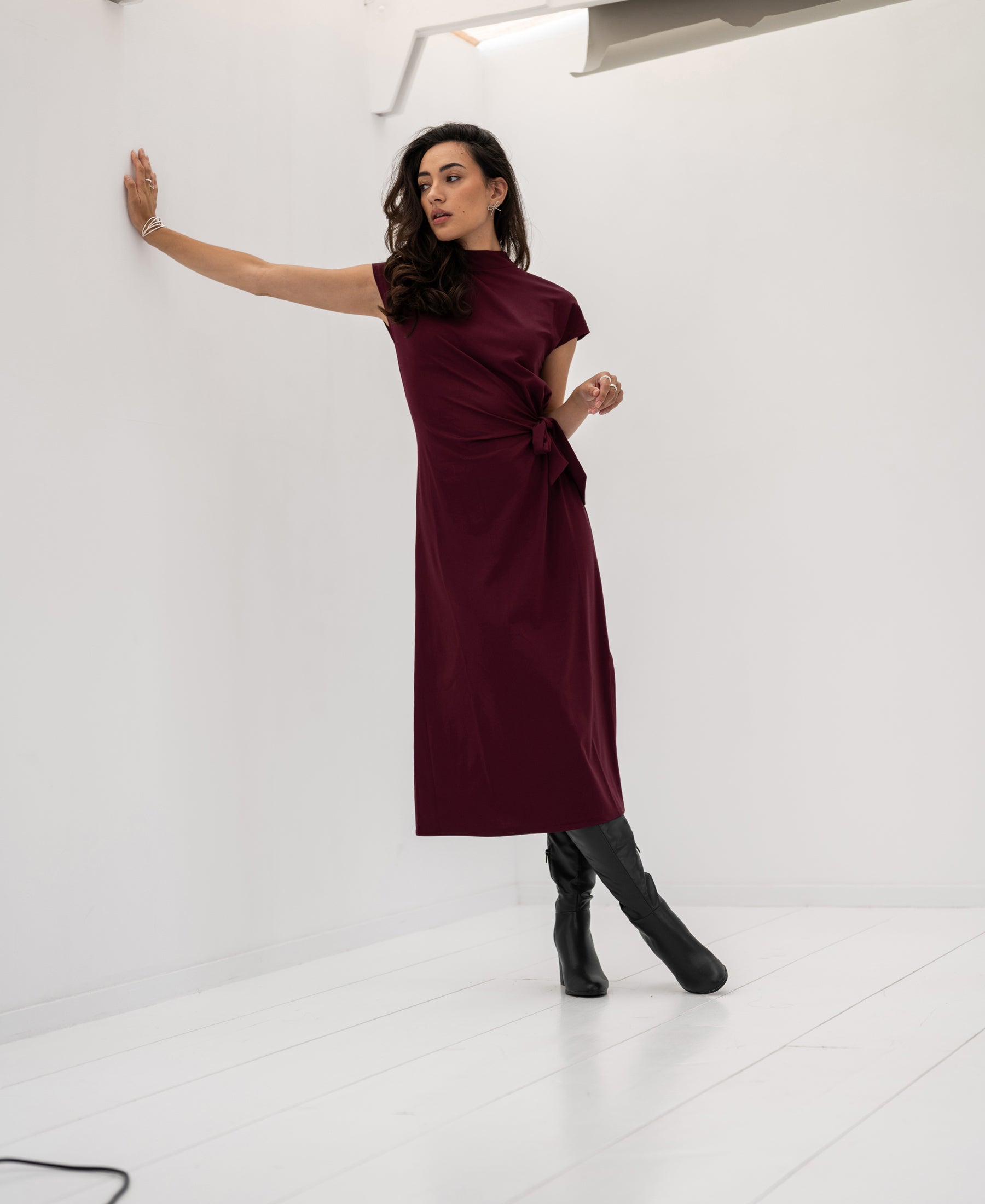 Woman wearing a Bordeaux PLEIN PUBLIQUE LA TOULOUSE travel dress stands against a white wall in a bright room.