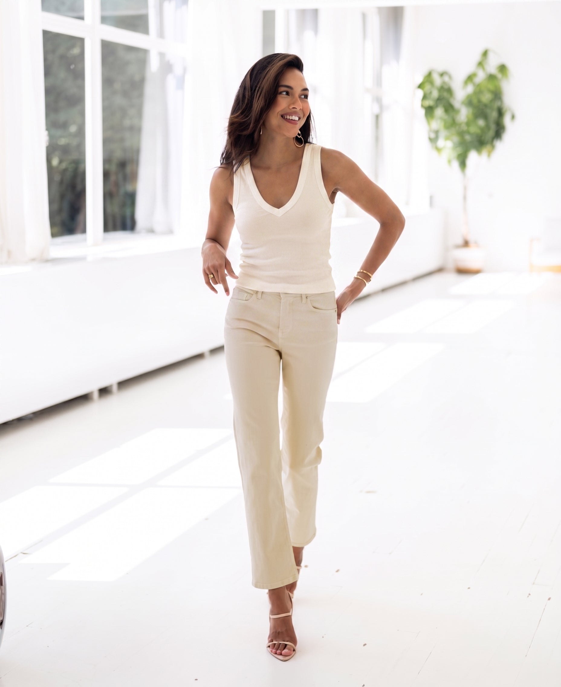 Woman wears soft butter-colored LA VALE top by PLEIN PUBLIQUE, standing in a sunlit room with large windows.