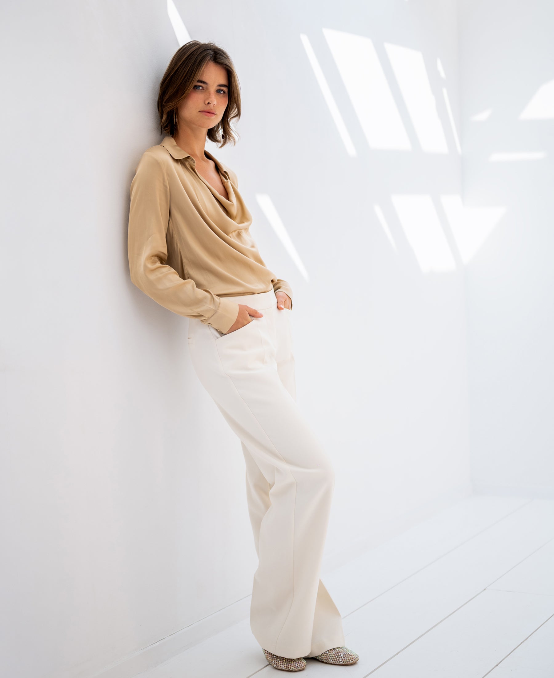 Woman wears the satin stretch blouse LE VENT in light sand by PLEIN PUBLIQUE, hands in pockets.