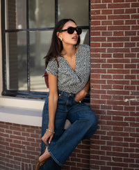 Woman wearing the PLEIN PUBLIQUE LA VERNIN Dot Charcoal polka dot top, leaning against a brick wall.