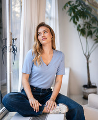 Light blue LA VERONIQUE V-neck T-shirt by PLEIN PUBLIQUE shown worn by a woman sitting cross-legged.