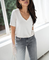 Woman wearing cream bamboo T-shirt stands by a sunlit window.