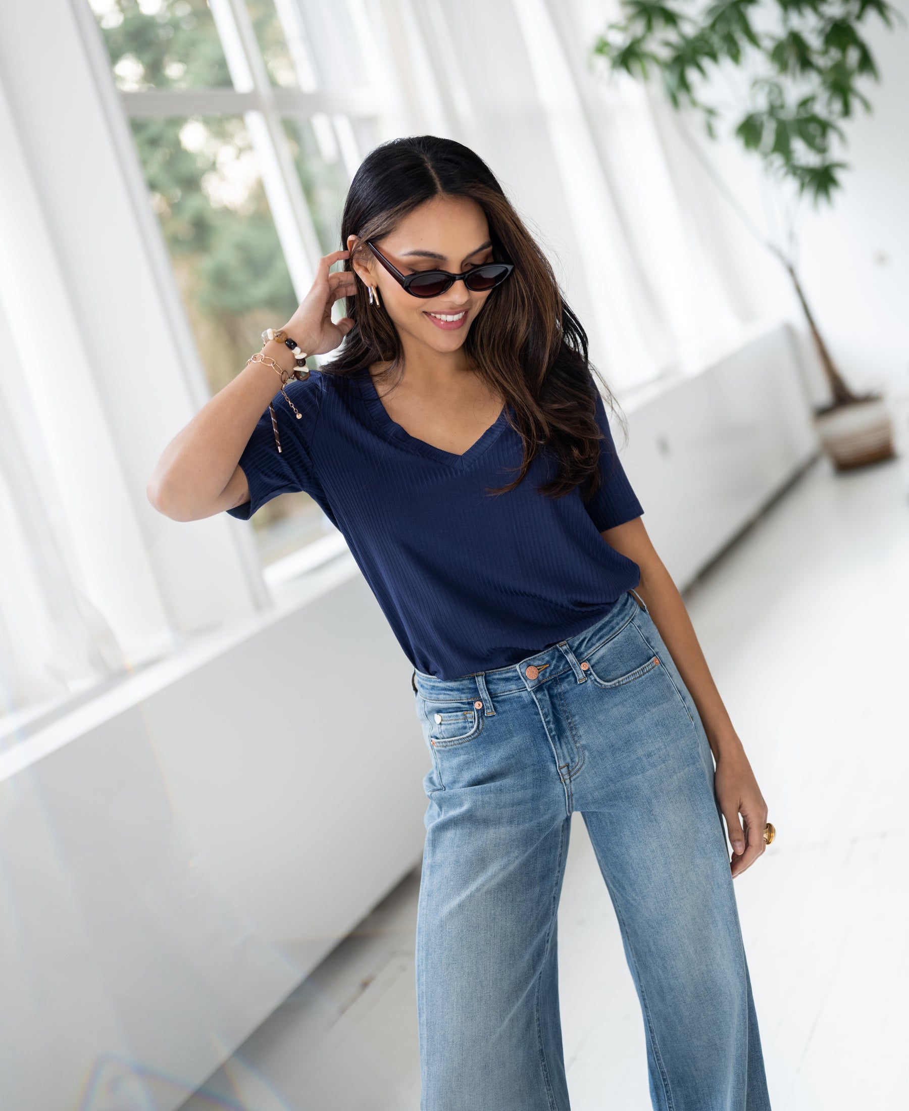 Smiling woman in sunglasses wears the PLEIN PUBLIQUE bamboo T-shirt in indigo by large windows.
