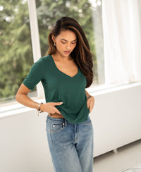 Woman wearing a green bamboo T-shirt by PLEIN PUBLIQUE stands by a window, adjusting her shirt.
