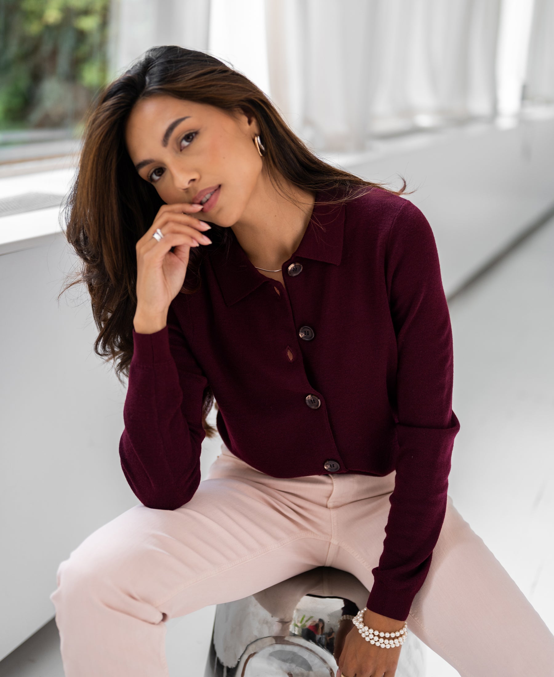 Woman wearing a Bordeaux red collared vest by PLEIN PUBLIQUE sits indoors, hand near her face.