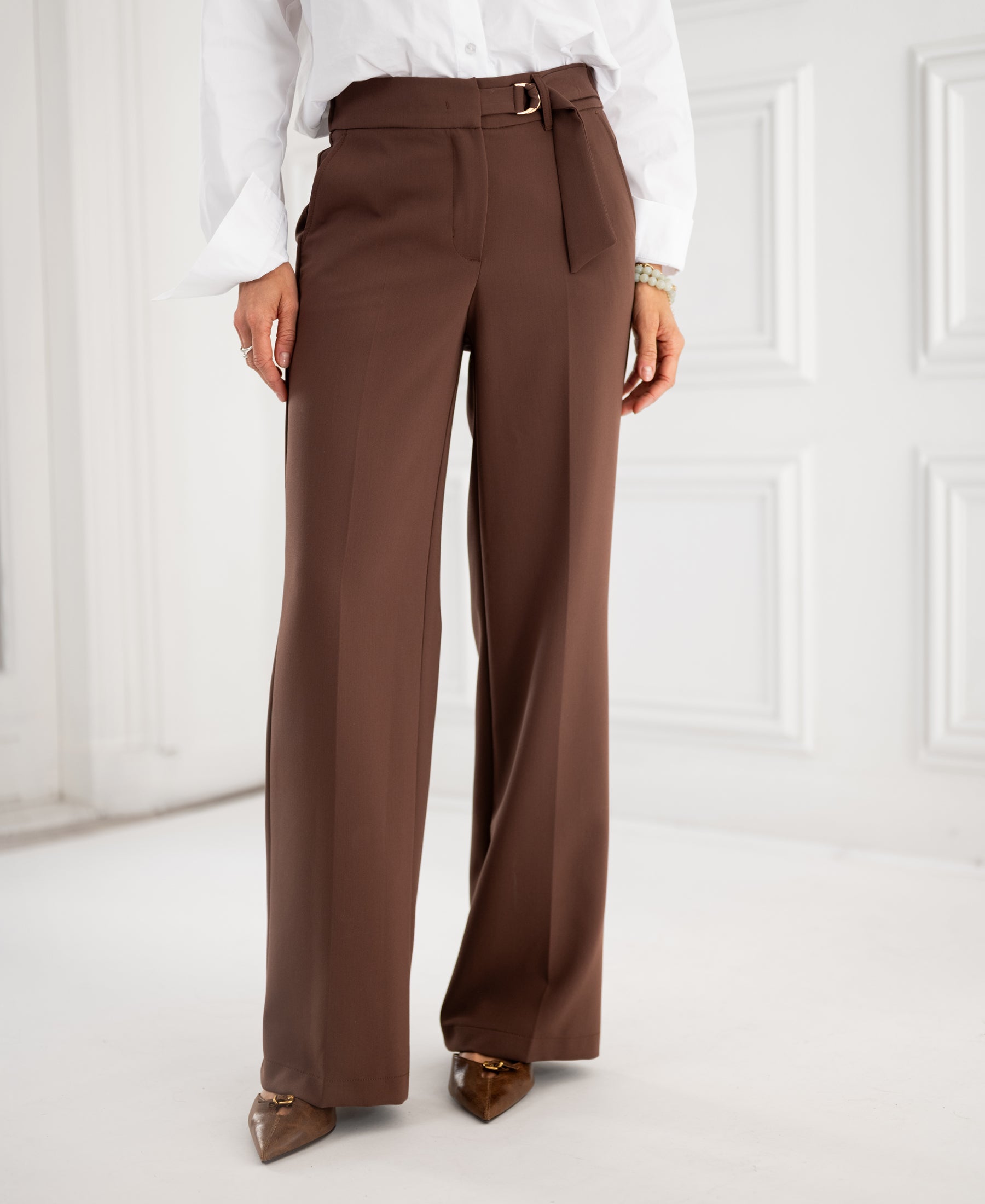Woman wears brown PLEIN PUBLIQUE's LA WENDE pants, standing indoors.