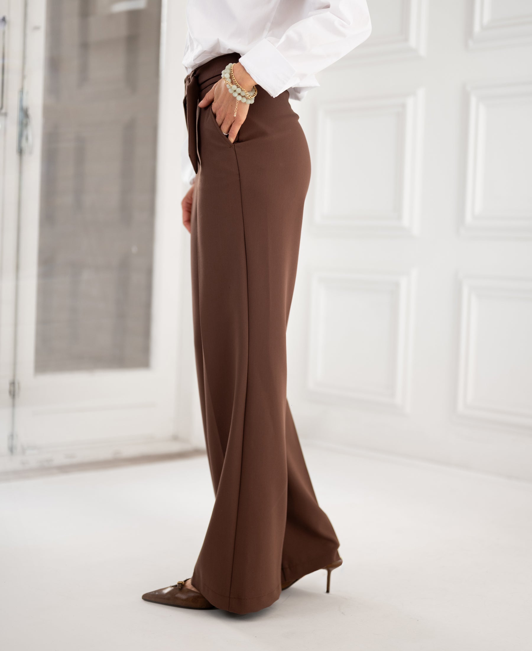 Woman wearing brown PLEIN PUBLIQUE LA WENDE pants in a minimal, bright white-walled room.