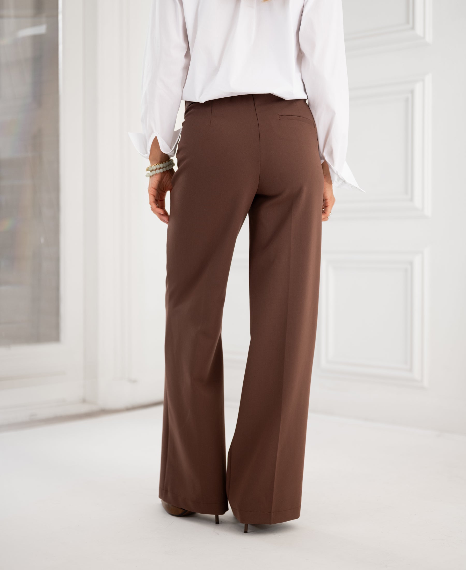 A person stands indoors facing away, wearing brown PLEIN PUBLIQUE LA WENDE trousers.