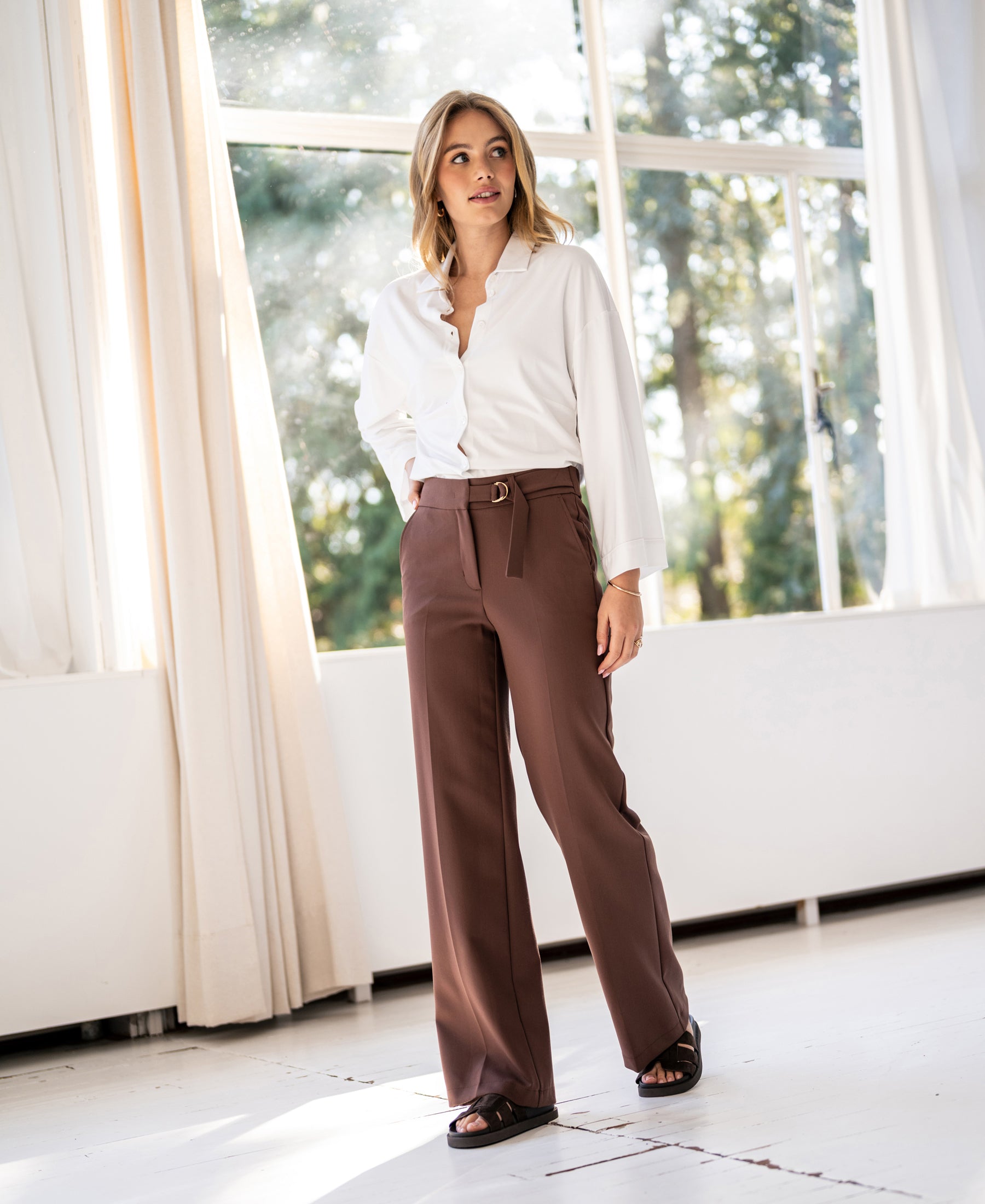 Woman wearing brown LA WENDE pants stands indoors by sunlit windows.