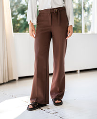 Person wears brown LA WENDE pants by PLEIN PUBLIQUE, standing by a window.