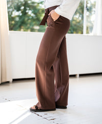A person wears the PLEIN PUBLIQUE LA WENDE Cacao trousers near a sunny window.