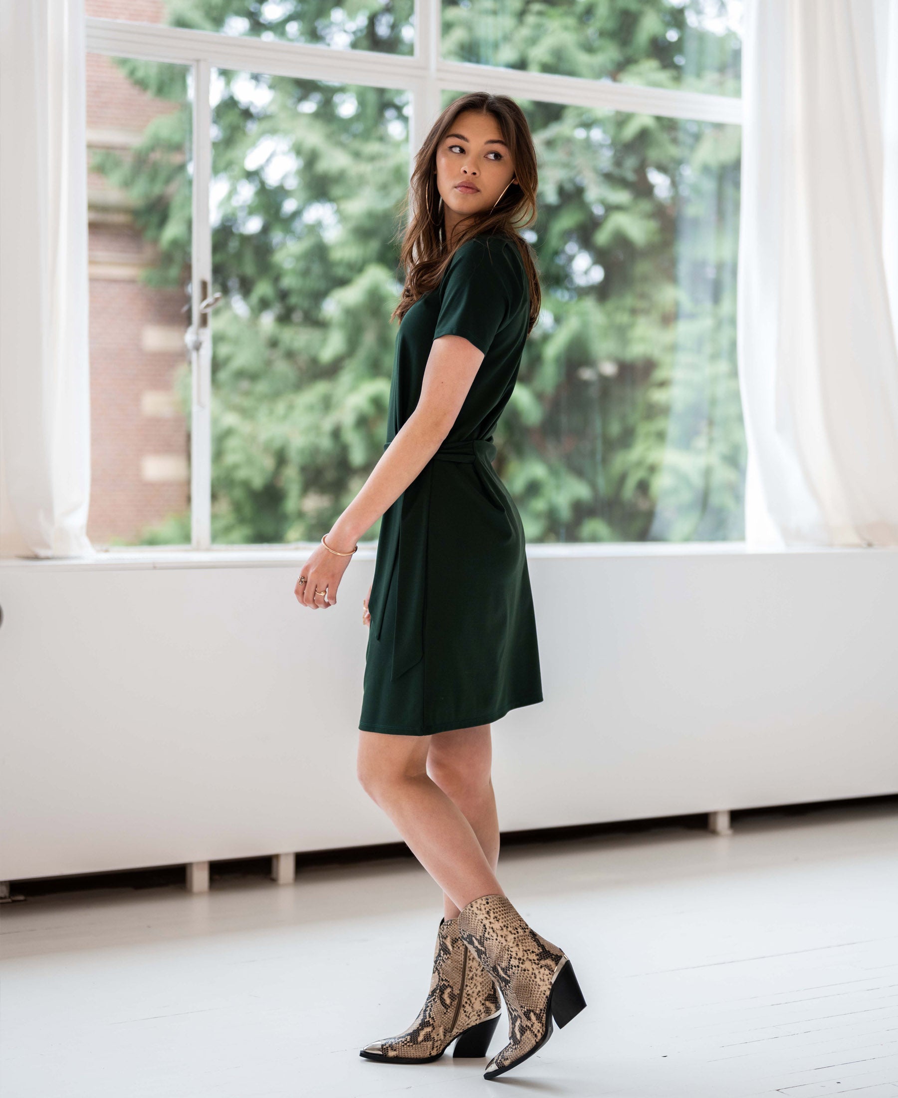 Woman wearing a dark green PLEIN PUBLIQUE button dress stands by large bright windows.